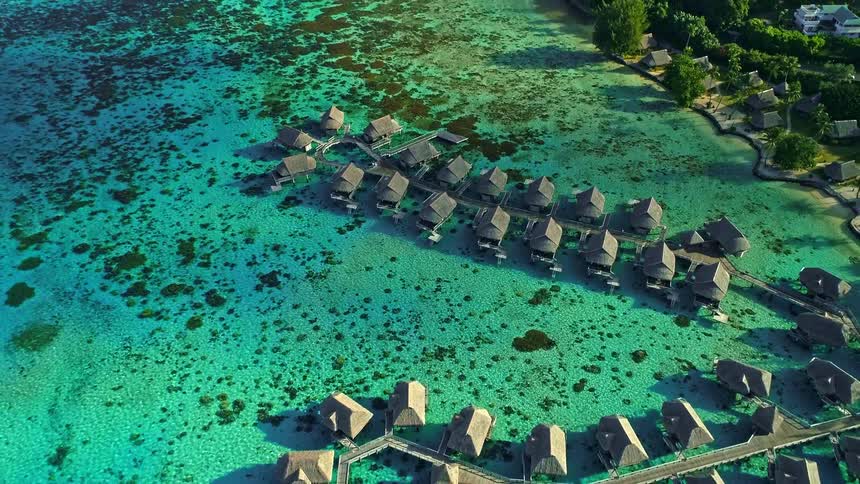 An incredible aerial top-down shot pulls back to reveal a stunning luxury resort of overwater bungalows in a pristine turquoise lagoon on a tropical island