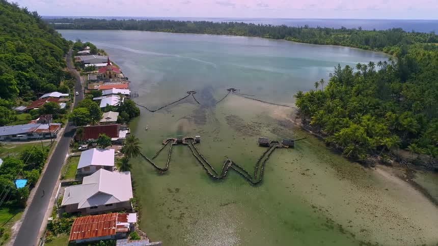 A slow aerial pull-back reveals ancient stone fish traps in a shallow, turquoise lagoon next to a picturesque tropical village