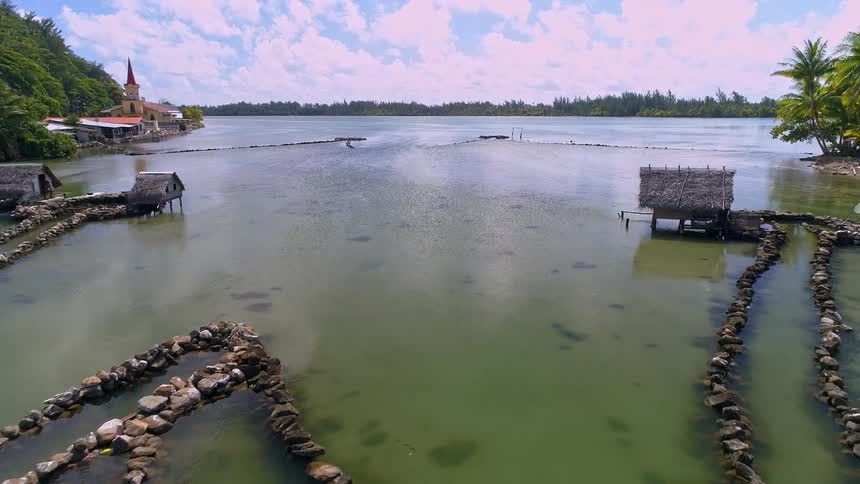 An aerial pullback shot reveals ancient stone fish traps and traditional thatched-roof huts in a tranquil tropical lagoon next to a village with a church