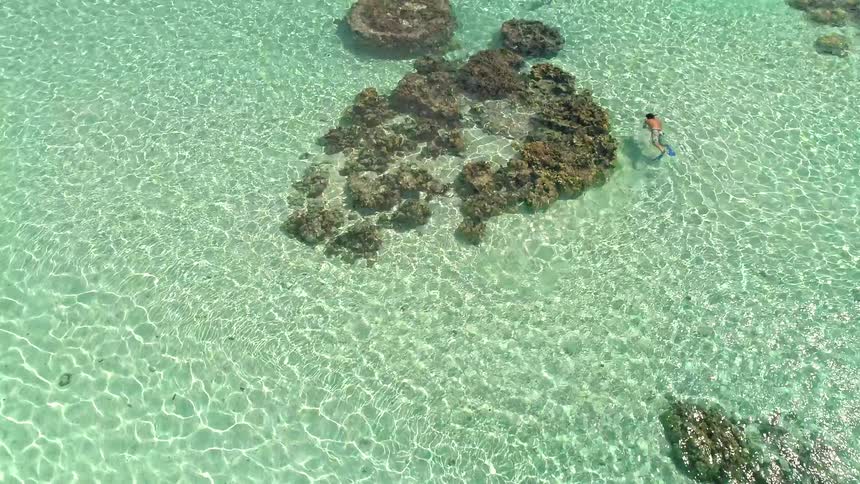 Aerial view starts on a snorkeler in clear turquoise water and pulls back to reveal a stunning tropical island with lush mountains