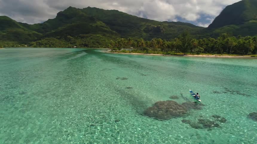 Stunning aerial footage circles a person paddling an outrigger canoe through the crystal-clear turquoise lagoon of a remote tropical island