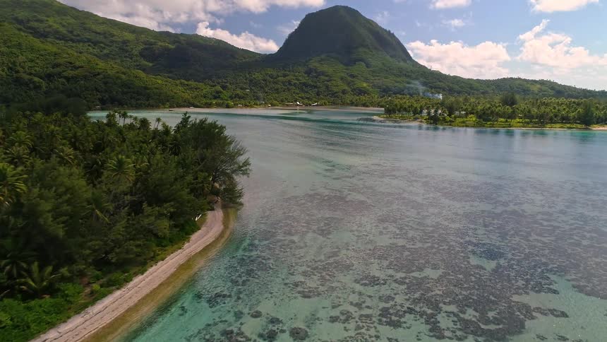 An incredible aerial view slowly flies over a pristine tropical island with a turquoise lagoon, sandy beach, and lush green mountains