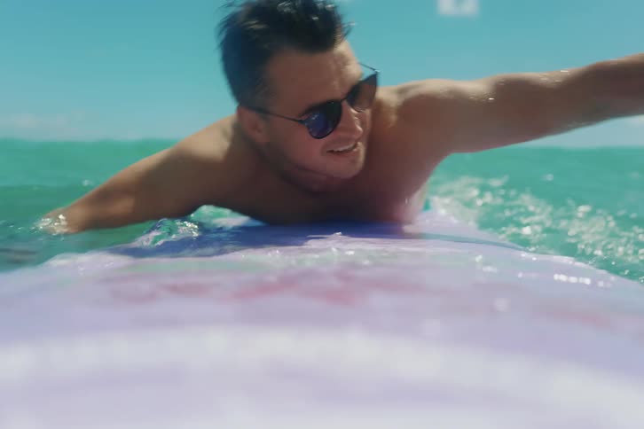 A smiling young man in sunglasses paddles on his surfboard in the clear blue ocean in a slow-motion POV shot