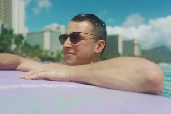 A smiling man wearing sunglasses relaxes on his surfboard in the tropical ocean with the Waikiki, Hawaii skyline in the background