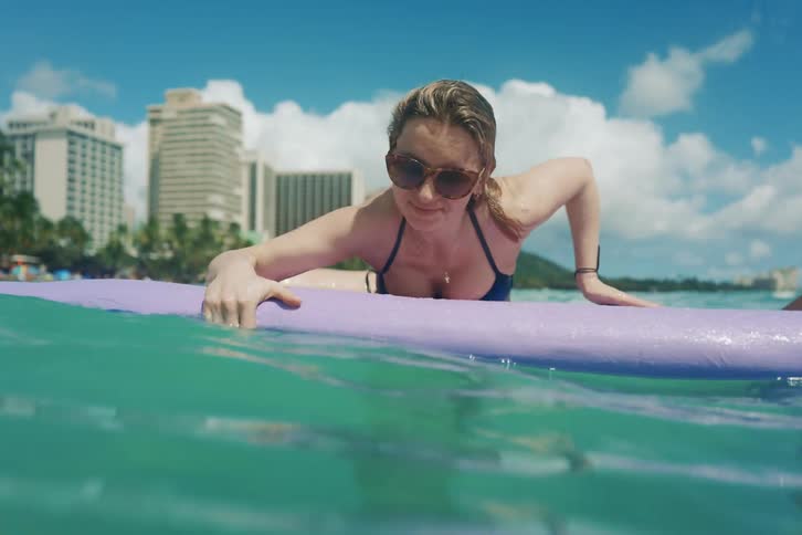 A young woman has fun trying to get on a surfboard with her boyfriend, but slips off into the tropical ocean water with a splash