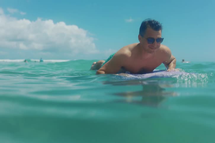 Happy surfer with sunglasses paddling on his board in the turquoise ocean on a beautiful summer day