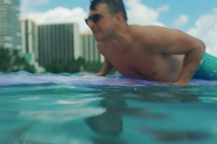 A man on a surfboard paddles in the ocean, then sits up and relaxes with a view of a tropical city skyline