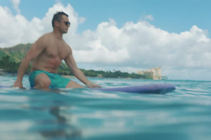 A male surfer puts on his sunglasses while relaxing on his surfboard in the calm tropical ocean