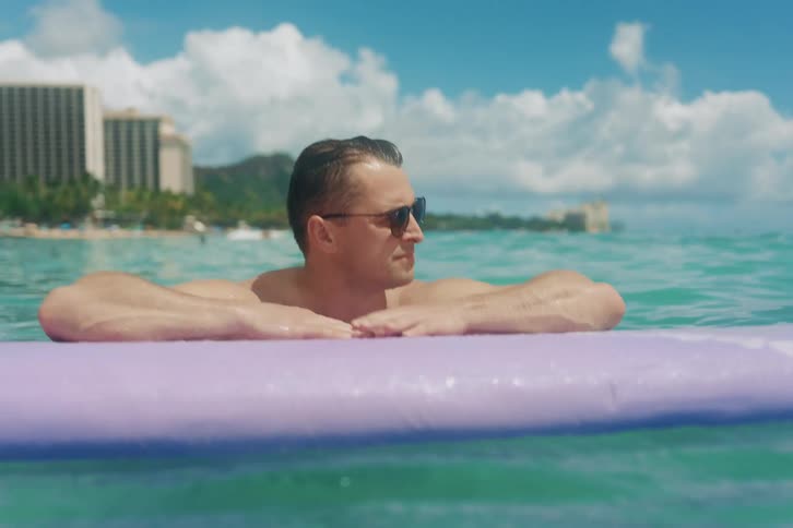 A man in sunglasses relaxes on a surfboard in the ocean, with Diamond Head and Waikiki hotels in the background