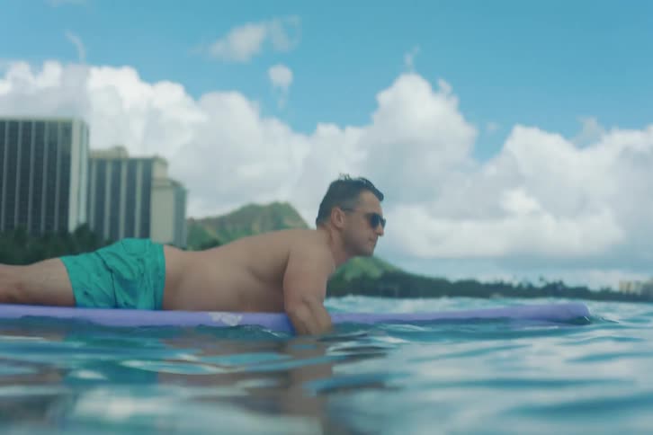 A man in sunglasses paddles on his surfboard in the calm ocean with the Waikiki, Hawaii skyline in the background
