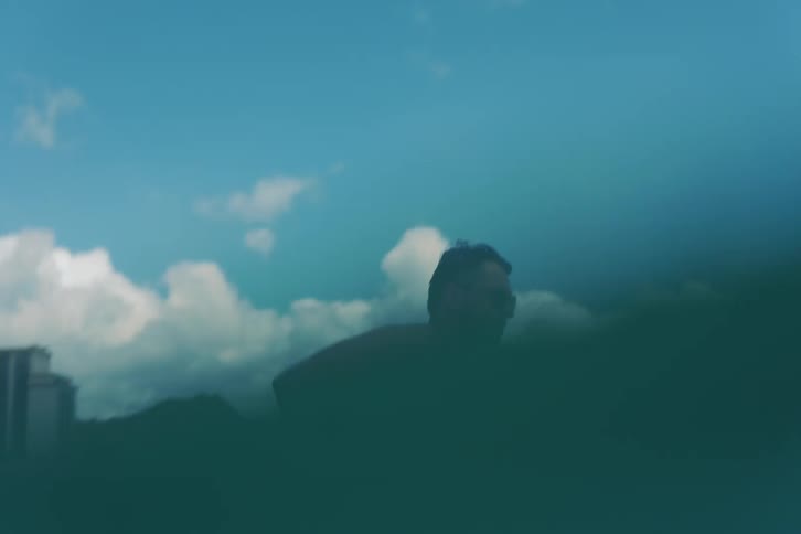 A man in sunglasses lies on his surfboard in the calm ocean, waiting for a wave with the Waikiki skyline in the background