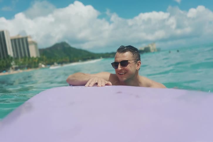 A smiling man in sunglasses relaxes on his purple surfboard, floating in the beautiful turquoise ocean on a sunny day