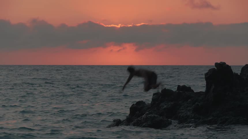 A man's silhouette is captured in slow motion as he heroically dives from a rocky cliff into the ocean during a vibrant sunset