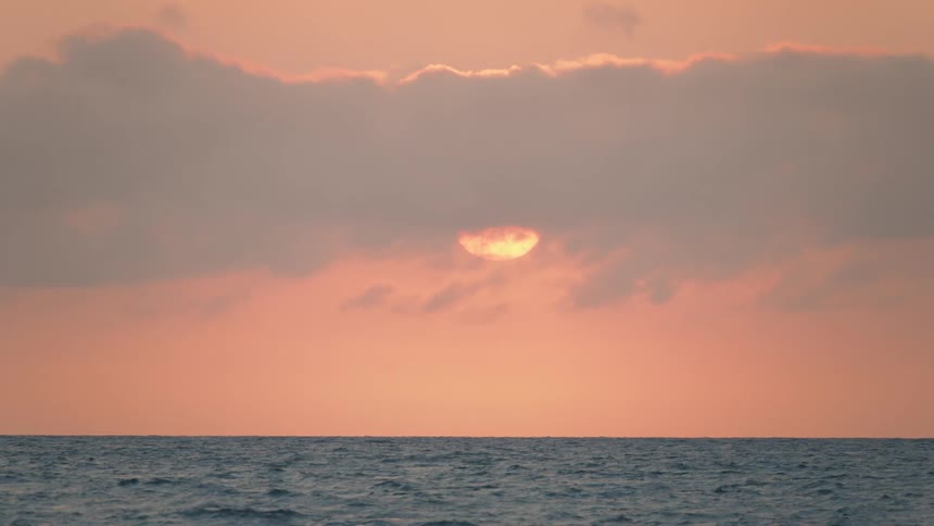 The sun glows behind a veil of clouds over the ocean, painting the sky a warm pink during a serene sunset