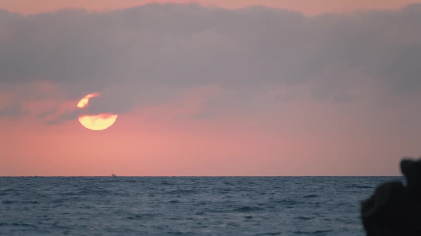 A serene view of the sun setting over the tranquil ocean, partially hidden by clouds as a distant boat sails across the horizon