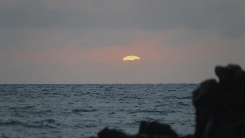 A time-lapse of a glowing yellow sun setting over the ocean horizon behind a veil of soft clouds