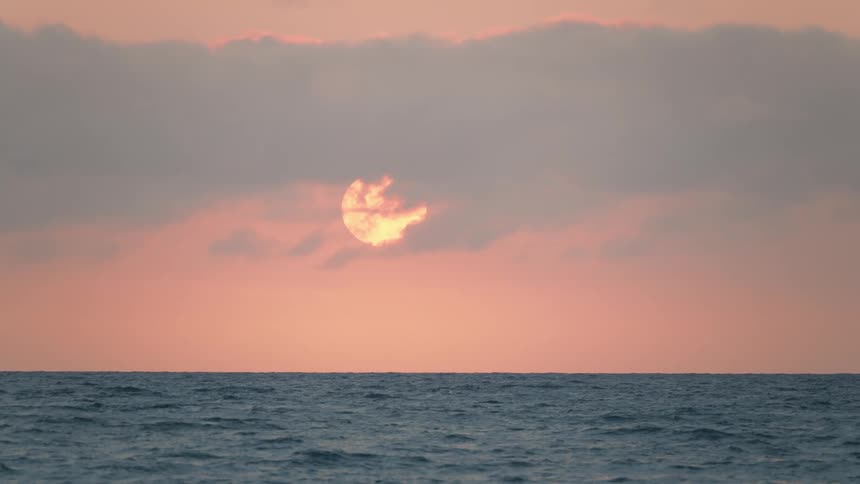 The sun slowly sets over the ocean, its golden light peeking through a layer of soft pink clouds