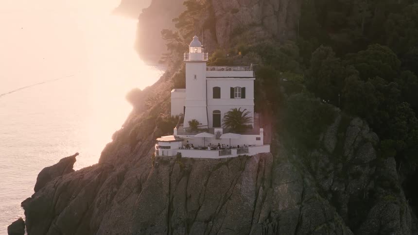 A stunning aerial view of the Portofino lighthouse perched on a rocky cliff, glowing in the warm, hazy light of sunset over the sea