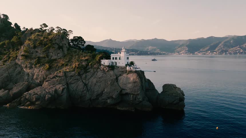 An aerial view slowly approaches the iconic white lighthouse of Portofino, perched on a rocky cliff overlooking the serene Italian Riviera at sunrise