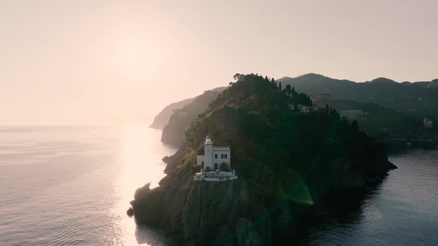 A cinematic aerial shot slowly pulls back to reveal a beautiful white lighthouse on a rocky, tree-covered cliffside during a golden sunrise