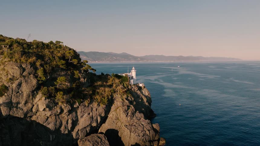 An aerial drone shot slowly approaches a picturesque lighthouse perched on a rocky cliff overlooking the beautiful Italian Riviera at sunrise