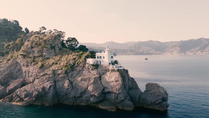 Cinematic aerial view reveals the iconic Portofino Lighthouse standing majestically on a rocky headland overlooking the Ligurian Sea