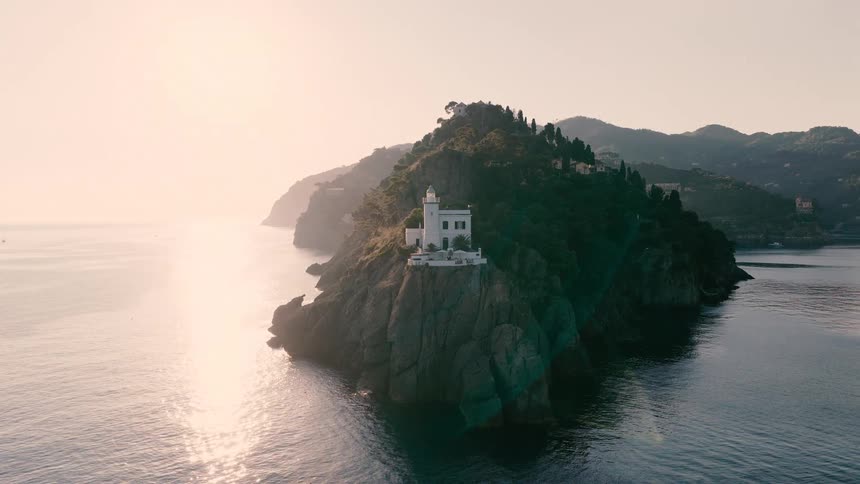 A stunning aerial pullback shot reveals a historic white lighthouse perched on a rocky, tree-covered peninsula along the Italian Riviera at sunrise