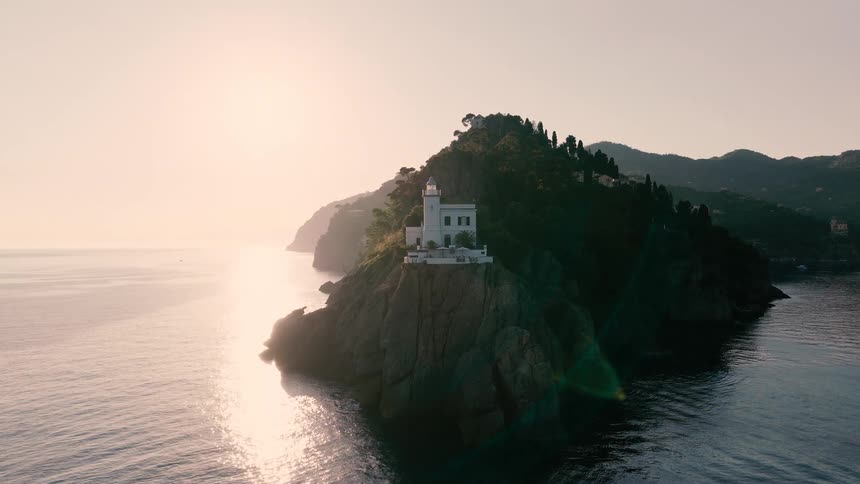 Aerial footage flying towards a beautiful white lighthouse on the rocky coast of Portofino, Italy, during a golden sunrise