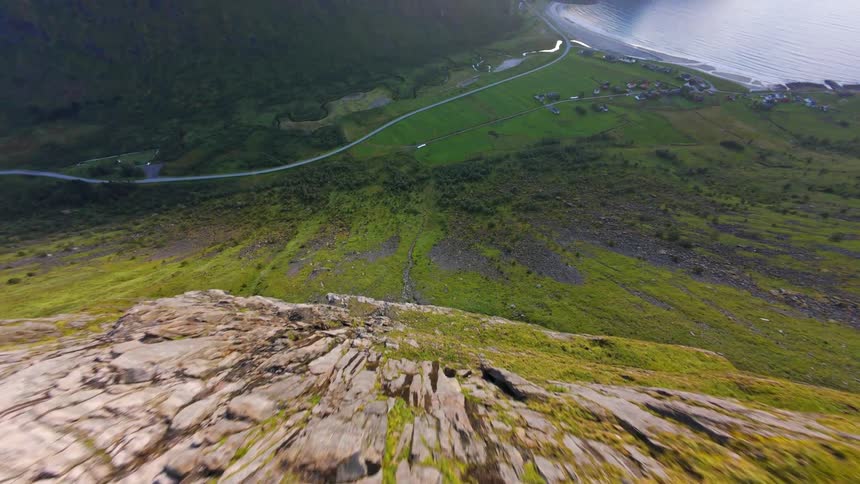 An exhilarating FPV drone shot swoops down a steep, rocky mountain face to reveal a beautiful coastal valley in Norway