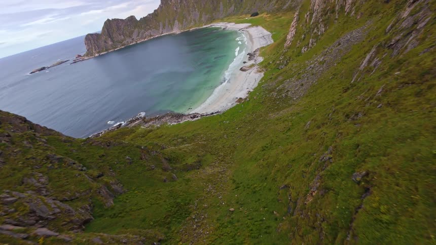 An FPV drone soars over a rugged mountain ridge to reveal a stunning hidden beach in a dramatic coastal cove