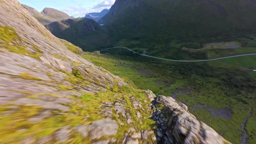 An exhilarating FPV drone flight dives down the side of a massive, rocky mountain revealing a lush green valley below
