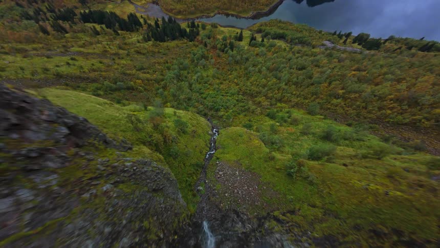 An exhilarating first-person view drone dive follows a cascading stream down a rugged, green mountainside, revealing a stunning Nordic valley and its tranquil lake