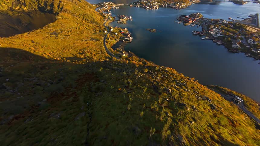 An exhilarating FPV drone dive down a sun-kissed, autumn-colored mountain ridge reveals a picturesque fishing village on the serene waters of a Norwegian fjord