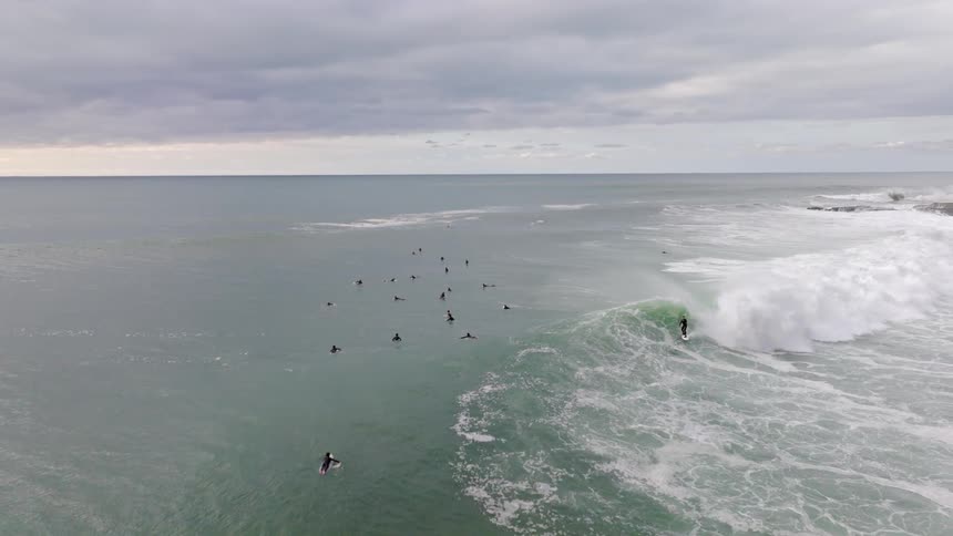 An aerial slow-motion shot of a group of surfers in the ocean as one catches and rides a large breaking wave under an overcast sky