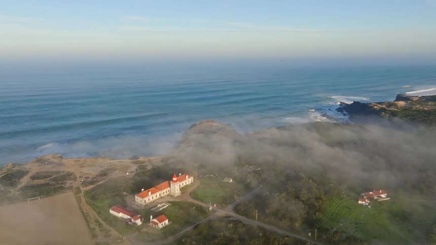 An aerial shot reveals a beautiful lighthouse on a foggy coastline overlooking the vast ocean with rolling waves