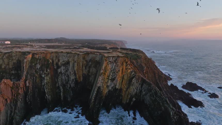 A slow aerial pullback reveals a stunning lighthouse perched atop dramatic cliffs along the coastline as birds fly over the crashing ocean waves at sunset