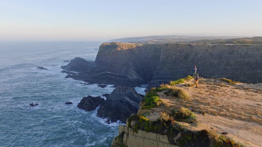 An inspiring aerial shot reveals two photographers on a cliff edge, capturing the magnificent view of the rugged coastline and crashing waves
