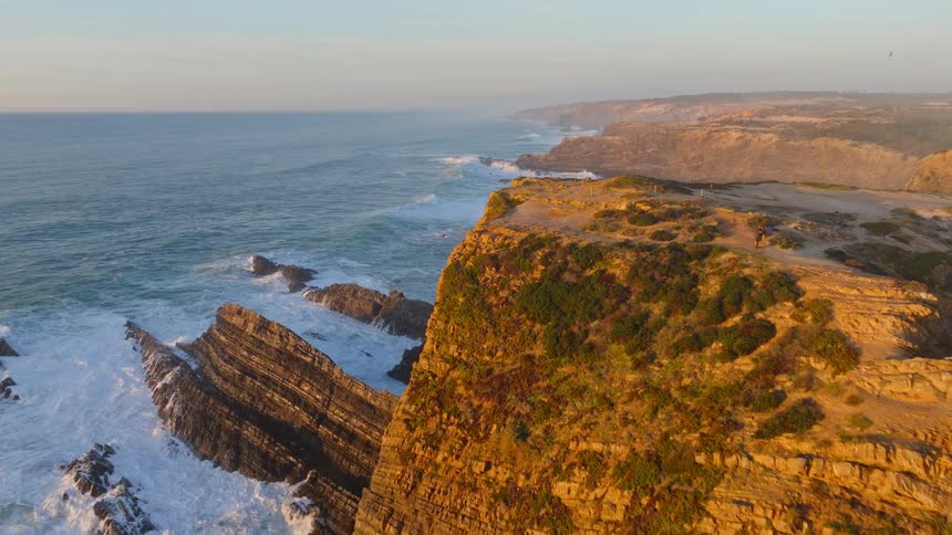 An epic aerial flight over rugged coastal cliffs reveals the dramatic shoreline as powerful waves crash below during a golden sunset