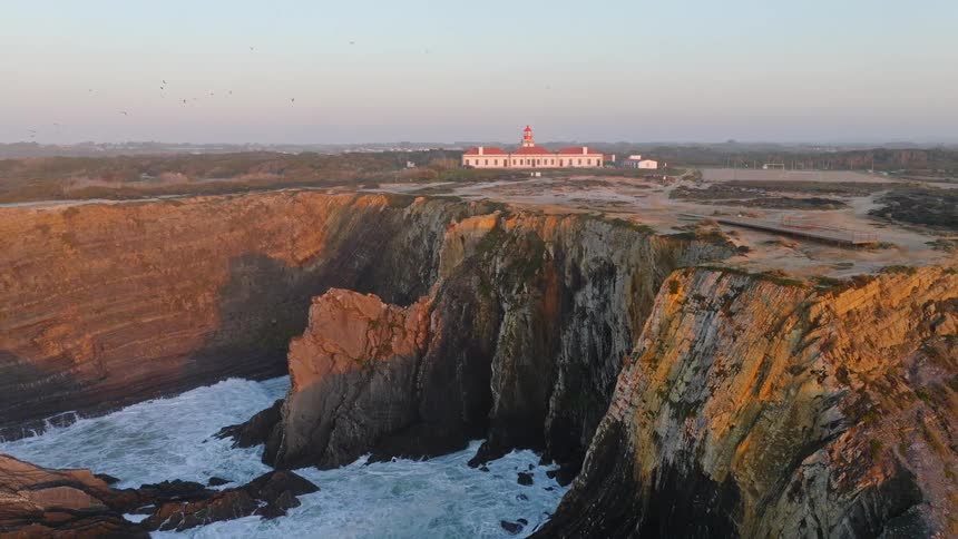 A majestic aerial pullback reveals the Cabo Sardao lighthouse perched on dramatic cliffs as golden sunlight illuminates the coastline and seagulls fly by