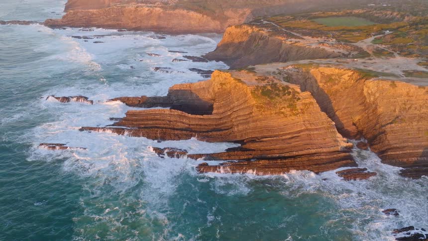 Aerial drone footage reveals a dramatic and rugged coastline as the camera pulls back from layered cliffs being hit by powerful ocean waves during a golden sunset