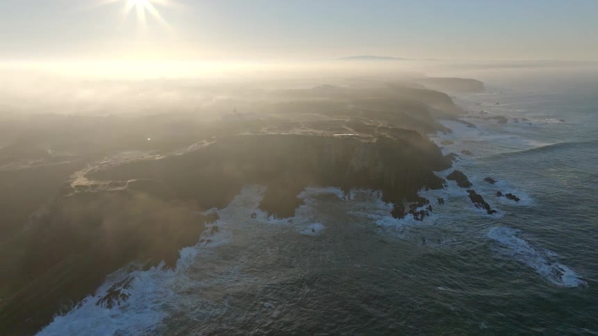 A stunning aerial view captures the sun shining through the mist over a rugged coastline and the expansive ocean