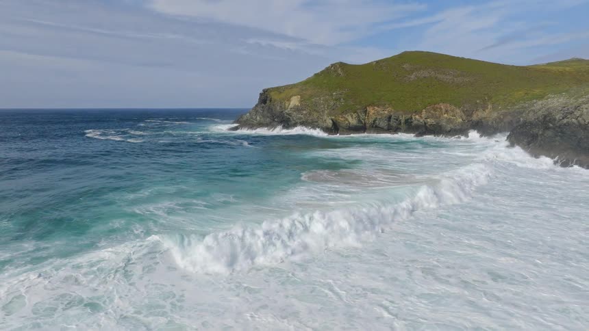 Turquoise ocean waves crash in slow motion against a rugged, green-covered headland on a sunny day