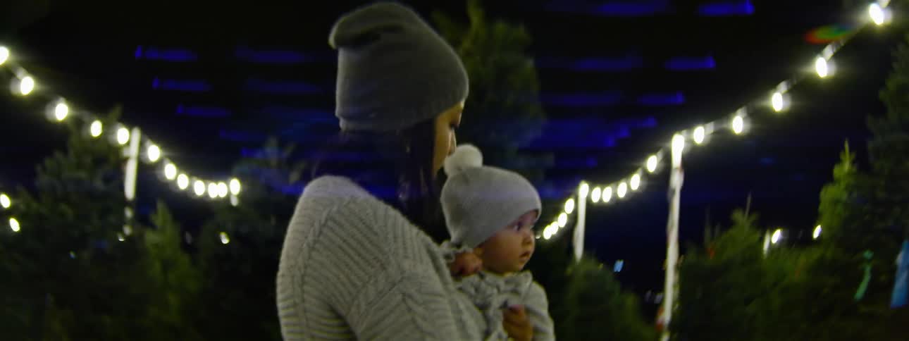 A woman holds a baby while browsing a christmas tree lot