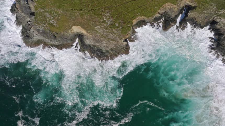 A mesmerizing top-down aerial slow-motion view of powerful turquoise ocean waves crashing against a rugged, grassy coastline