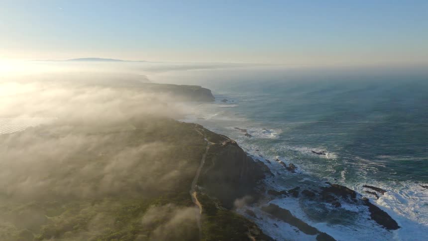 A cinematic aerial view of a foggy coastline with waves crashing against the rocky cliffs during a beautiful sunrise