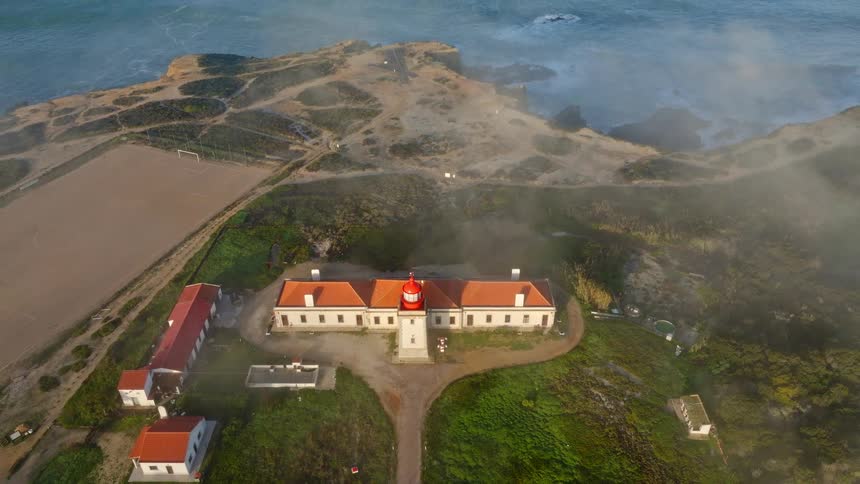 A beautiful aerial view rises from a top-down shot of a lighthouse on a foggy cliffside overlooking the ocean