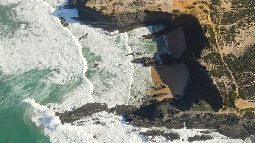 A static top-down aerial view captures powerful turquoise waves crashing against a rugged, rocky coastline and a secluded sandy cove
