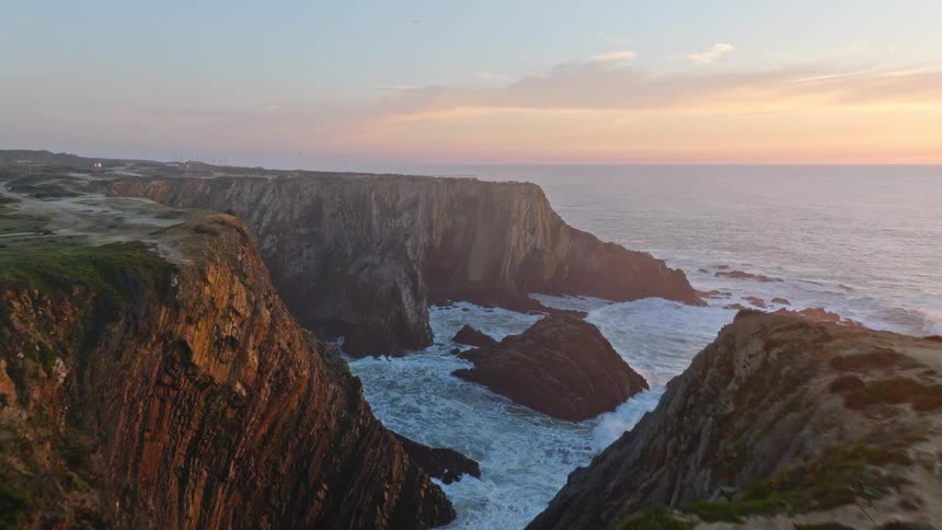 A stunning aerial view flies along a dramatic rocky coastline with a lighthouse as the sun sets over the ocean