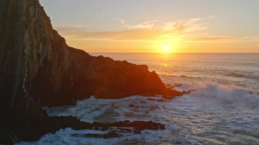An aerial view reveals a stunning golden sunset over a dramatic rocky coastline as powerful waves crash against the shore