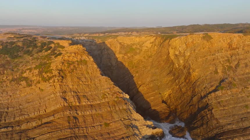 An aerial shot pulls back to reveal dramatic, layered cliffs on a rugged coastline bathed in the golden light of sunset